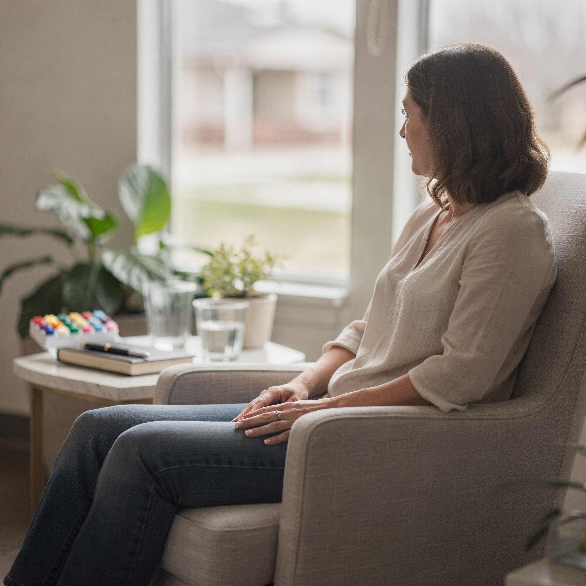 Woman sitting in a light-filled counseling room with plants and a notebook on a side table